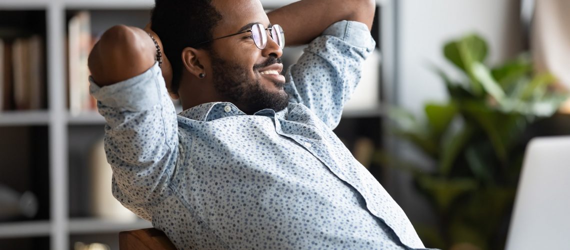 Relaxed calm young african businessman resting looking away sit at desk with laptop hands behind head, satisfied office employee take break feel stress relief peace of mind concept chill at work